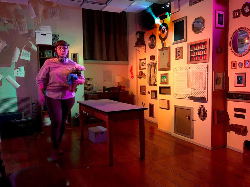 A woman performer, wearing a button down shirt and pants, carries a blooming potted plant. She walks in a room with archive boxes making up the ceiling, paper documents hanging in the corner, and a table and stools in the center. A wall made of archive boxes in on one side, and a wall on the other side is covered with framed images, drawers, cabinets, mirrors and slots.