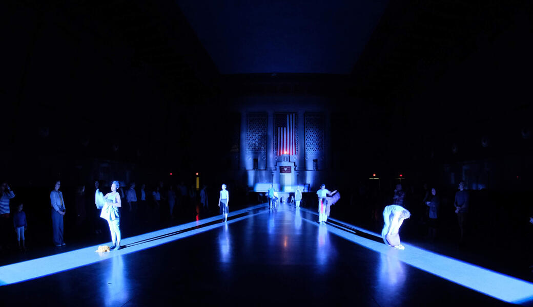 Performers stand in two shafts of light traveling in opposite directions across a large marble floor.