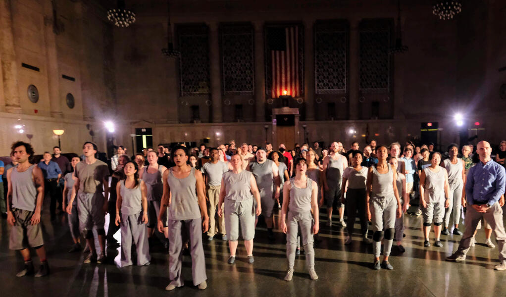 A large number of performers, all dressed in gray, stand still on a large marble floor and gaze upward in an expansive room.