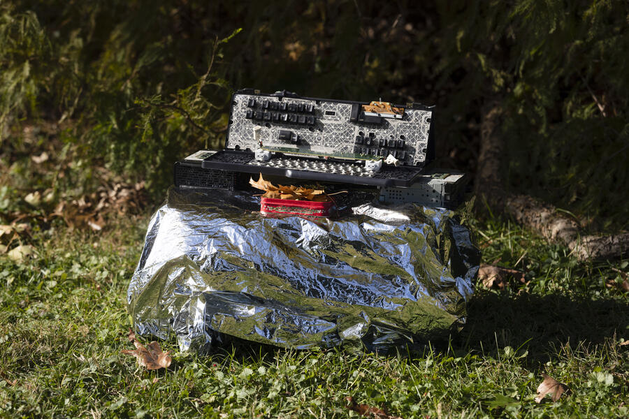 A sculpture made of discarded keyboards, keys and other computer components sits on a silver box in a grassy area at the foot of a tree.