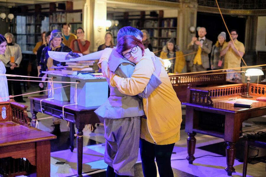 Two women performers, wearing costumes of different eras, embrace in a reading room in a library. Other performers, audience members, shelving full of books and gold ribbons criss-crossing the space are in the background.