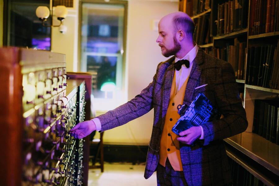 A male performer, wearing a bowtie, gold vest and tweed blazer and holding a clipboard, reaches for a drawer in a card catalog in a library.