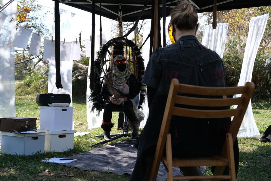 A performer in a futuristic costume sits in a swing chair inside a tent structure set in a park. Another performer is seated across from the first and the two of them are looking at each other.