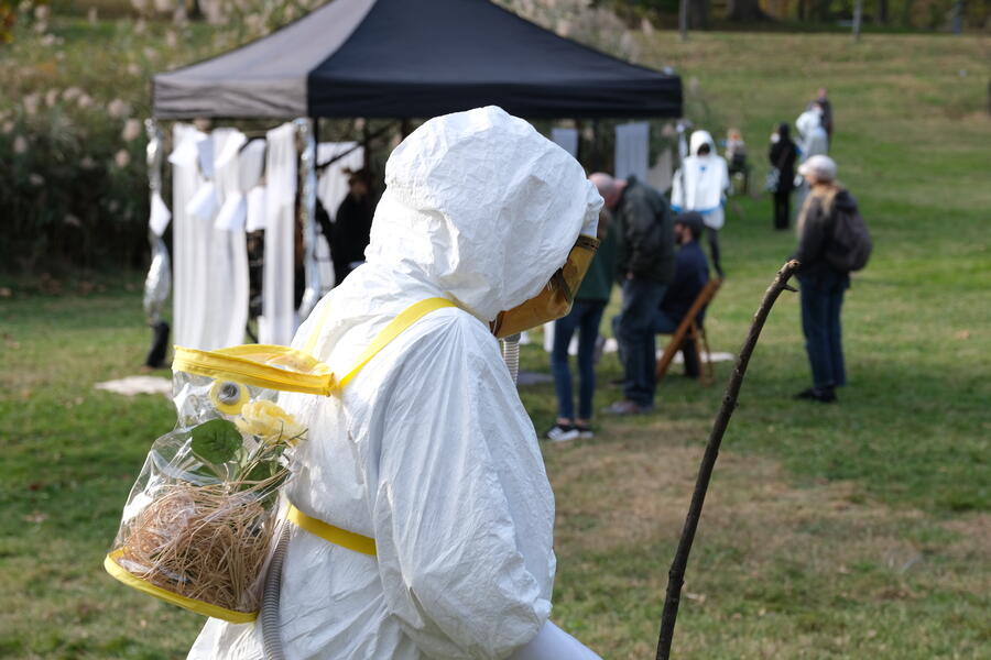 A performer dressed in a futuristic white hazmat costume and face shield walks through a grassy field in a park. A tent structure, audience members and trees are in the background.