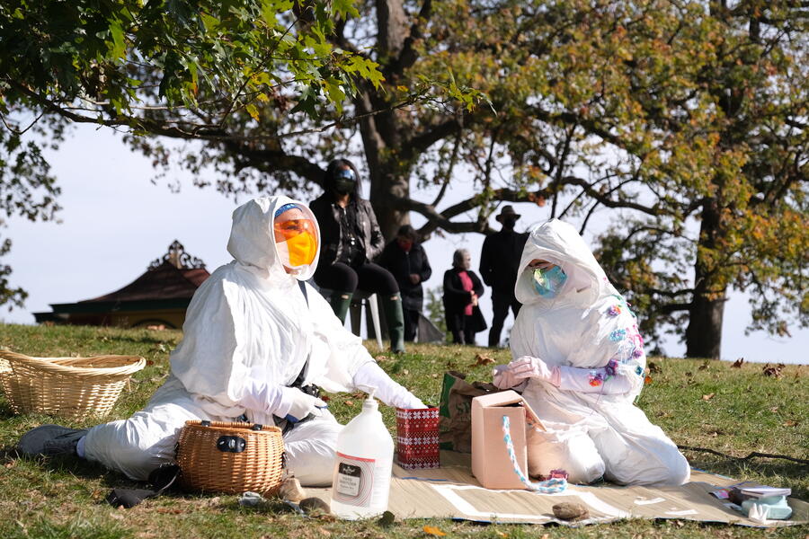 Two performers dressed in futuristic white hazmat costumes and face shields sit on a blanket on a grassy hillside. There is a basket and a variety of objects placed on the blanket. A large tree and audience members, standing, are in the background.