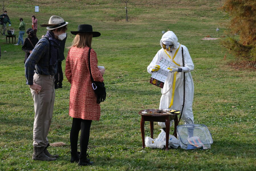 A performer dressed in a futuristic white hazmat costume and face shield unfolds a map onto a table set in a grassy field in a park. Two audience members look on.