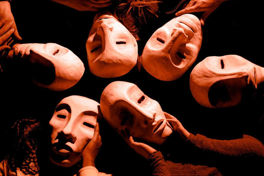 Six performers, all wearing white masks, lay in a circle on a marble floor with their heads touching.