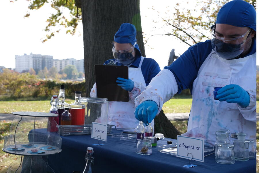 Two performers dressed in blue with white aprons and latex gloves stand behind a table covered with bottles, jars, tongs, and other items. One performer is writing on a clipboard. The table is set at the base of a tree in a park.