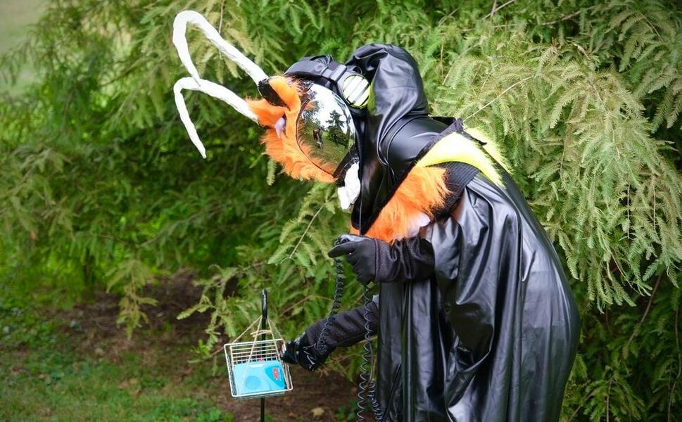  A performer dressed in a futuristic black hazmat costume and a face shield that looks like an insect reaches for a small tape player. The tape player is hanging in a wire basket at the base of a tree in a park.