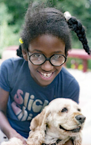 A photograph of Pamela Woolford as a grade-school child with her hair in three braids, wearing glasses, and smiling while kneeling behind her dog.