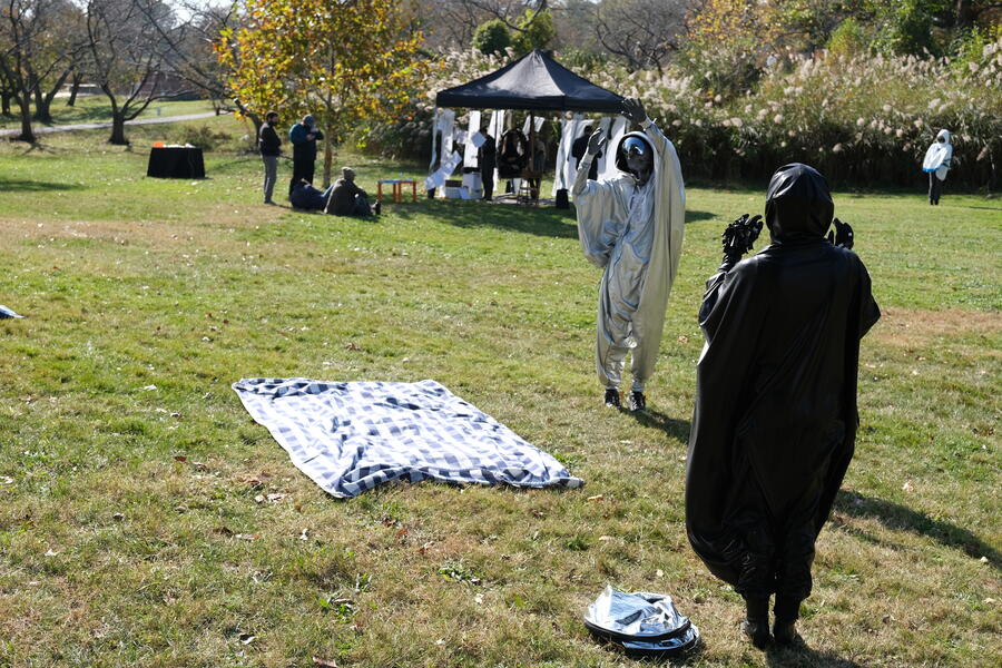 Two performers in "sack suits" interact at a distance in a park with a tent in the far distance.
