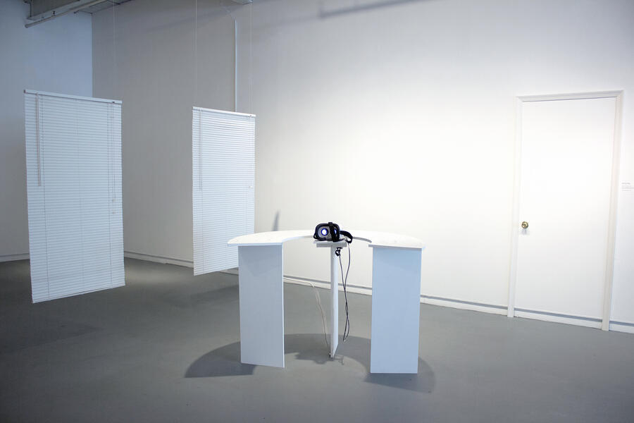 White gallery space with two blinds hanging in the back left, a white table with VR headset in the center, and a door frame to the right