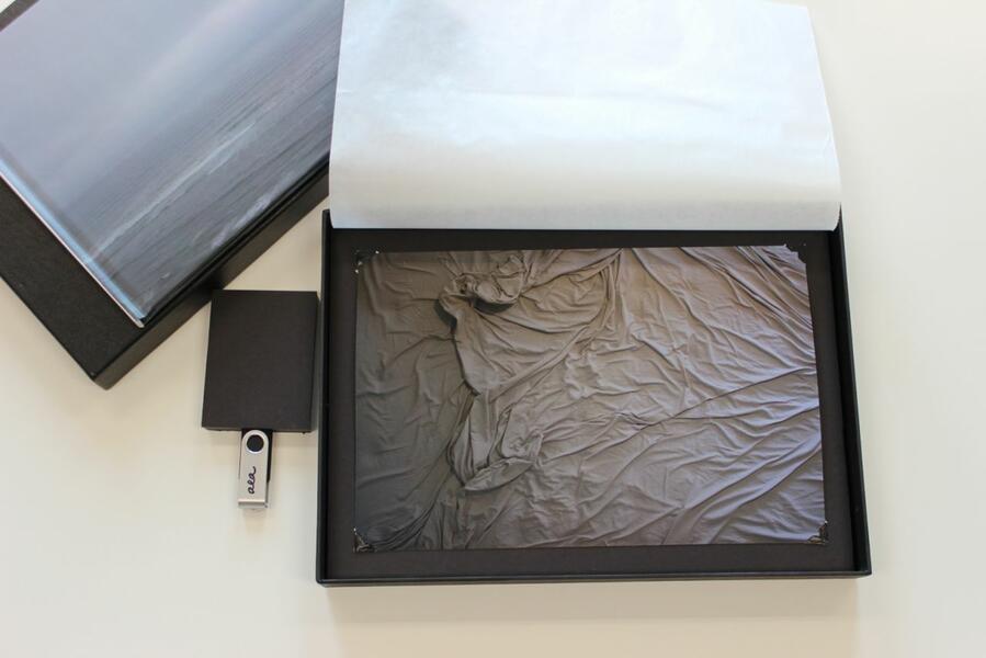 Photograph of gray bed sheets inside of a black box with protective film flipped open and flash drive beside the box