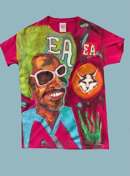 Photo of a hot pink tee, hand painted with a Black man with a green fro, a white and black cat face, and E A painted twice on it.